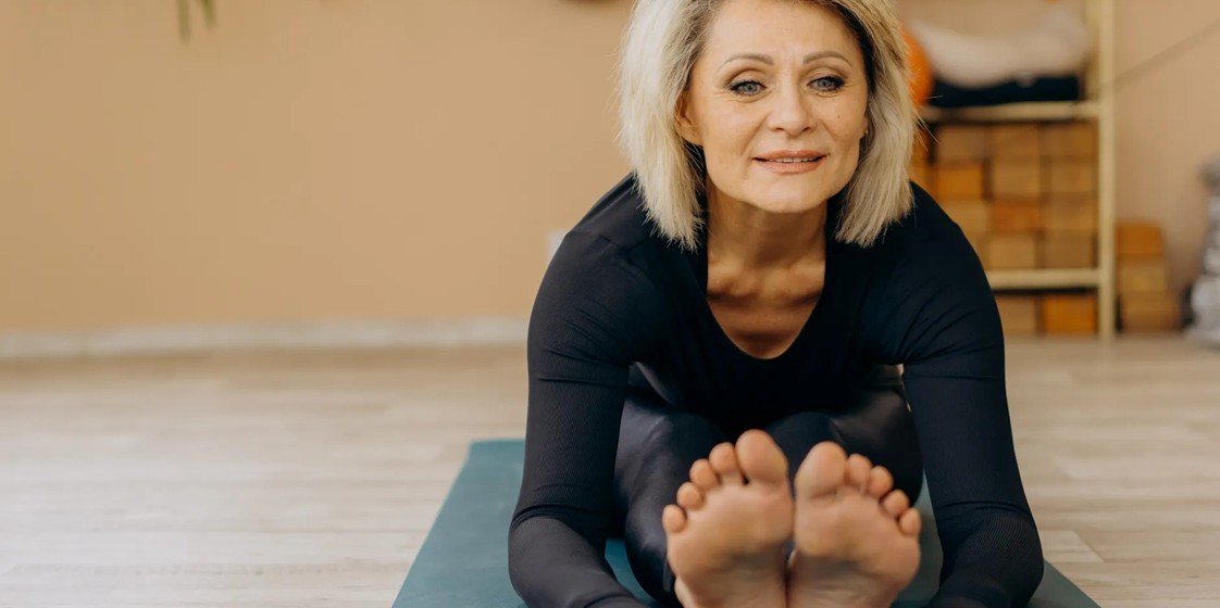 Mujer de más de 40 años sonriendo mientras practica un estiramiento de yoga sentada sobre su esterilla